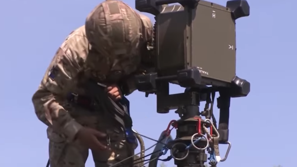 A soldier in camouflage gear operating an advanced electronic warfare device mounted on a tripod under a clear sky.