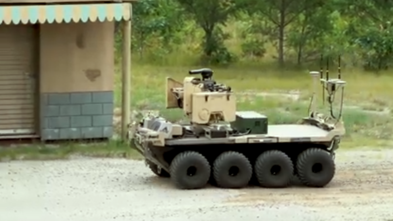 Eight-wheeled unmanned ground vehicle with a mounted turret moving on a gravel path near a small building and trees.