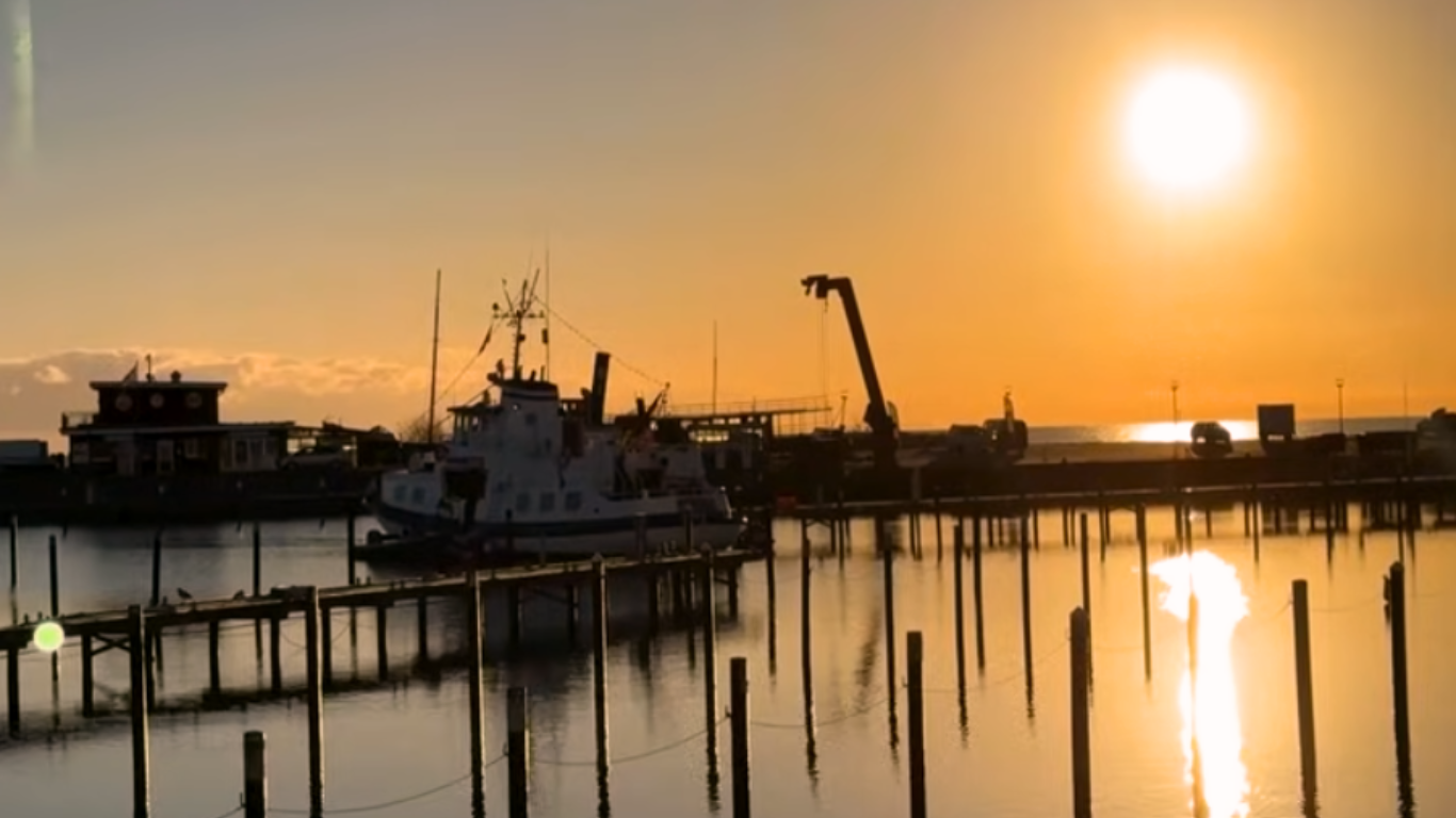 Harbor at sunset with docked vessel, pier structures, and crane silhouetted against calm water