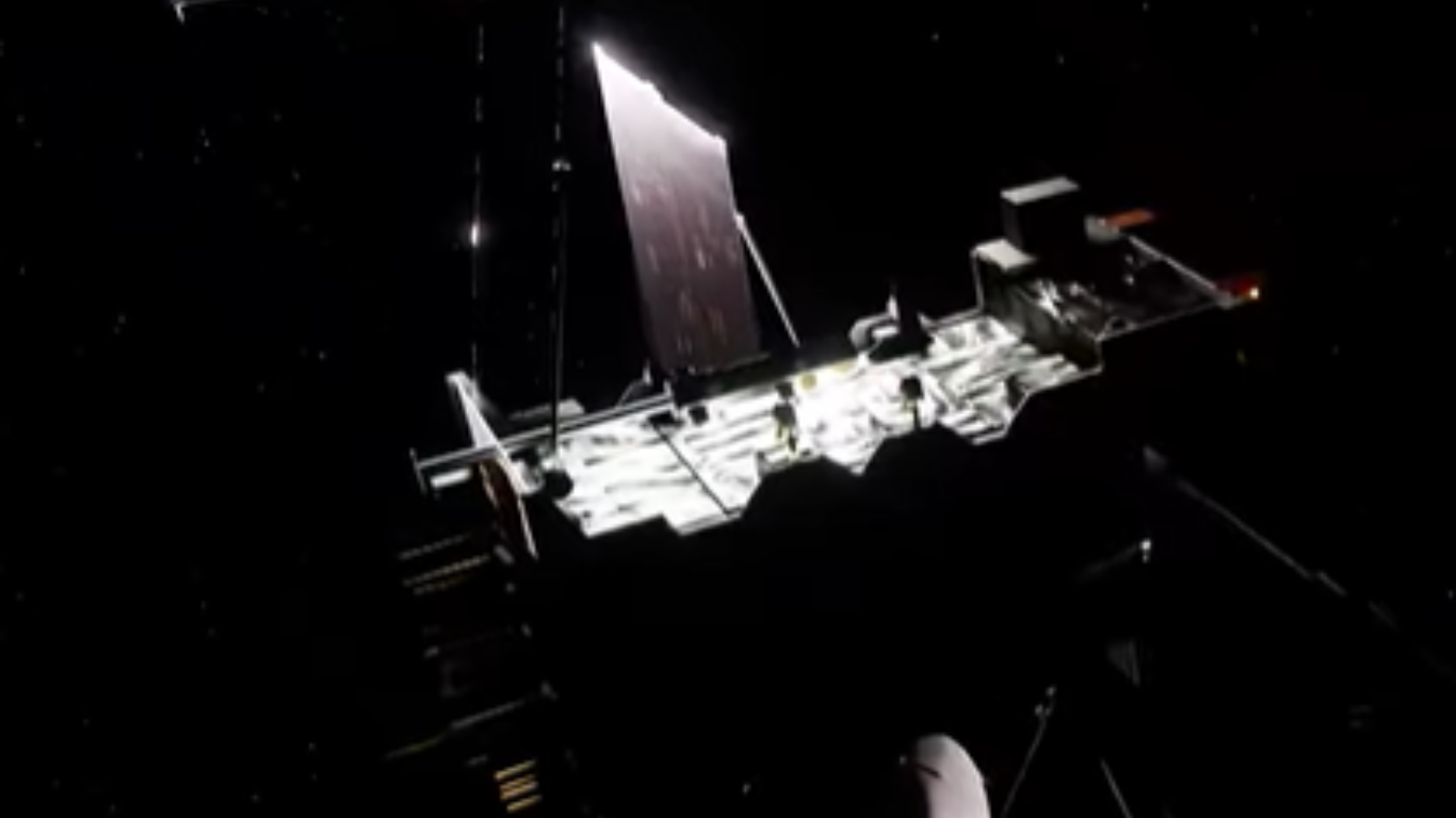 A spacecraft floating in deep space, illuminated against a dark star-filled background, with a large reflective panel or sail extending upward and structural components visible along its body.