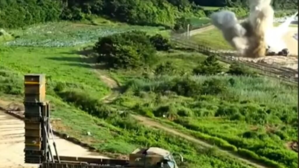 A truck-mounted vertical missile launcher deployed in a field near an explosion, linked to an AI combat cloud network.