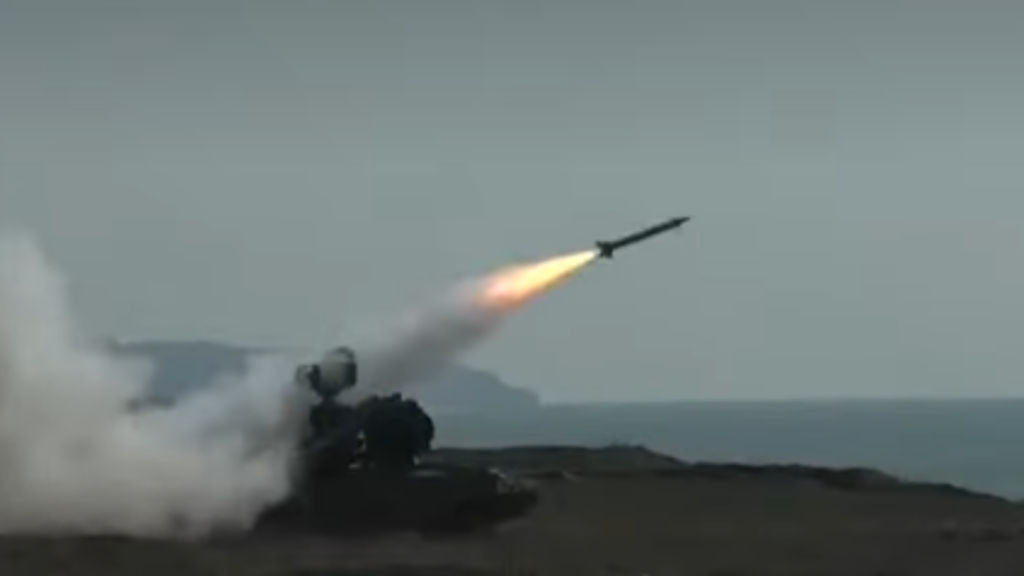 A surface-to-air missile launching from a mobile launcher by the sea, connected to an active AI combat cloud network.