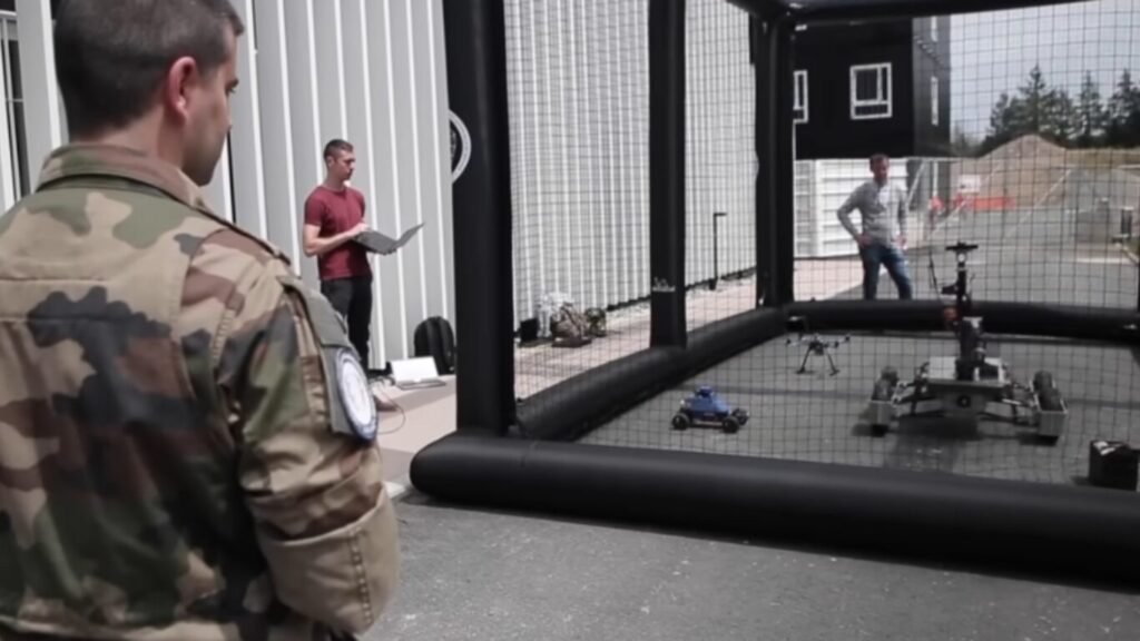 Military and civilian technicians testing unmanned rovers and aerial drones inside a safety netting enclosure, demonstrating field testing for AI Defence Operations.
