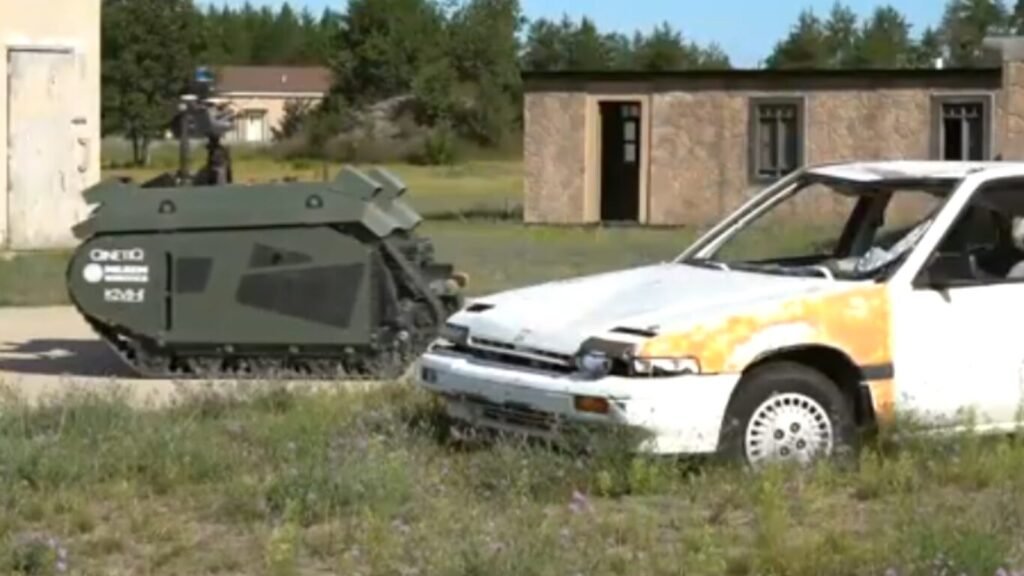 An unmanned, tracked robotic vehicle navigating past a white car in a simulated urban training village, demonstrating technology for AI Last Mile Resupply.