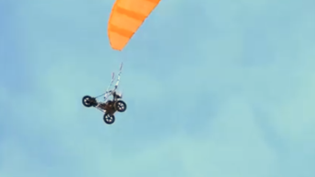 An unmanned paramotor or flying ATV vehicle suspended beneath a bright orange parachute canopy in mid-air, demonstrating aerial tech for AI Last Mile Resupply.