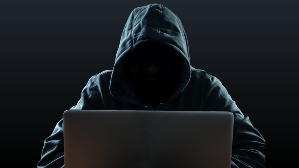An anonymous individual wearing a dark hoodie sitting behind an open laptop screen against a solid black background, illustrating the threat of AI-powered phishing.