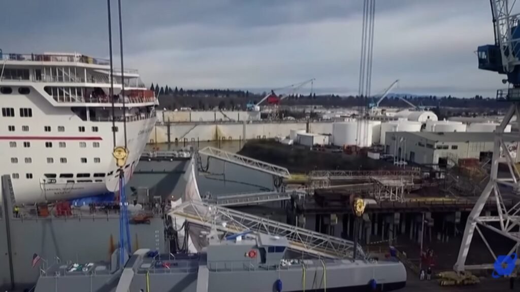 An aerial view of a military naval vessel being hoisted by heavy shipyard cranes next to a cruise ship, illustrating maritime platform staging for AI Warfare.