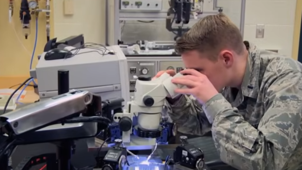 An Air Force technician in camouflage uniform looking through a laboratory microscope while working on Battlefield Robot Cybersecurity hardware components.