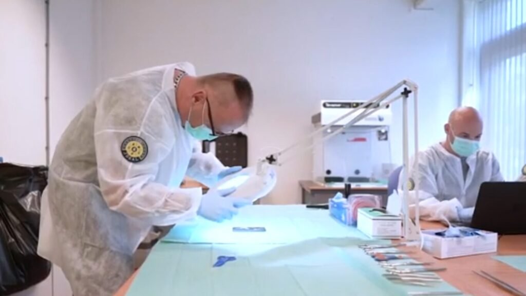 Forensic specialists in white protective gear inspecting evidence under a magnifying lamp in a lab, demonstrating analysis for Biometric IFF identification systems.