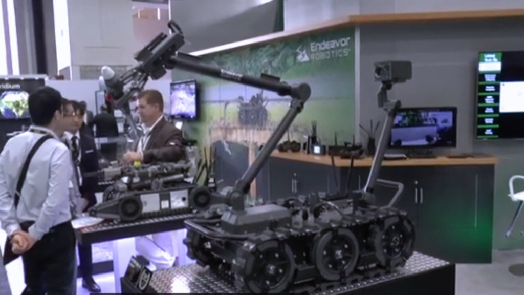 A military ground robot with tracks and a mechanical arm displayed at a technology convention booth showcasing Centaur Warfare capabilities to attendees.