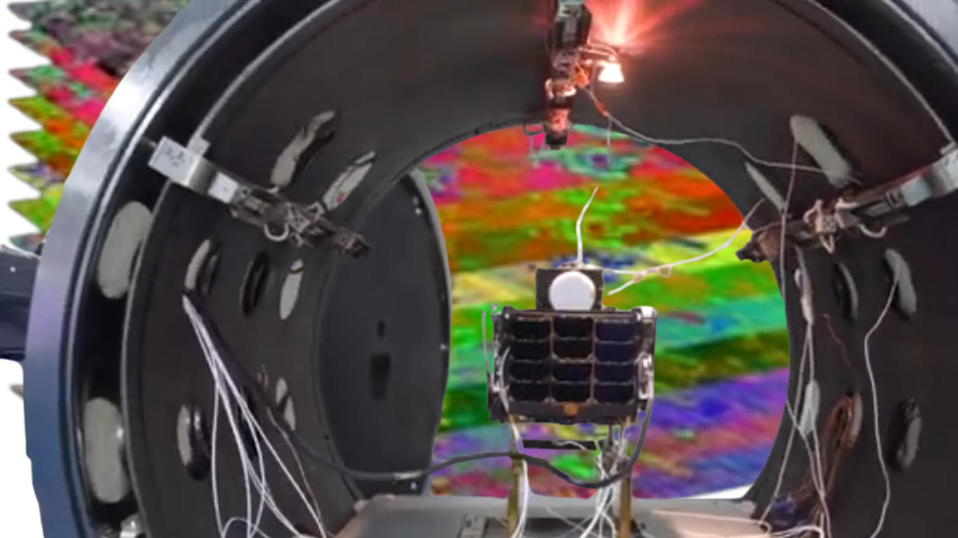 A small satellite-like device inside a cylindrical testing chamber, with wires attached and a colorful hyperspectral image displayed in the background.