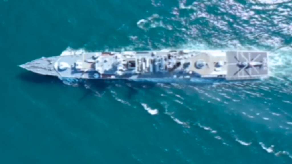 An overhead aerial view of a gray warship cruising through teal ocean waters with a helicopter parked on the flight deck, demonstrating operations linked to Military Robotics Naval Military Robots.