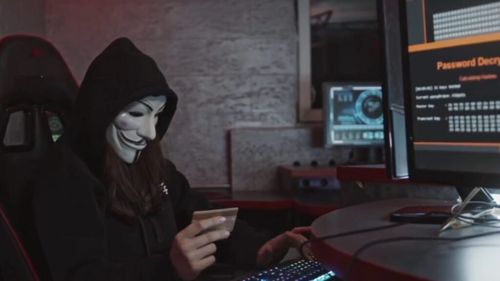An individual wearing a Guy Fawkes mask and a black hoodie sitting at a computer workstation holding a payment card, illustrating cybercrime threats like Satellite Hacking.