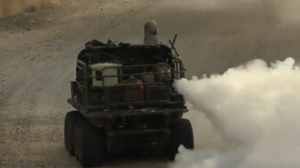 An unmanned military vehicle categorized under Stealth Ground Drones deploying a dense white smoke screen while driving on a dirt road.