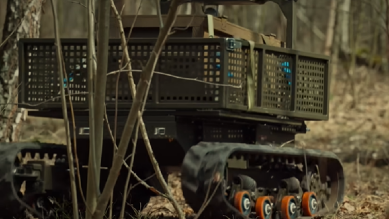 Tracked ground robot carrying equipment moving through a forest with dry leaves.