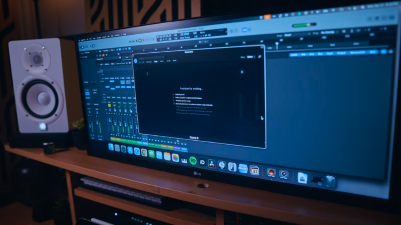 A wide-screen LG monitor displaying a digital audio workstation with an iZotope Ozone 9 Master Assistant window open, illustrating an automated AI Music Production setup on a wooden desk next to a white studio monitor speaker.