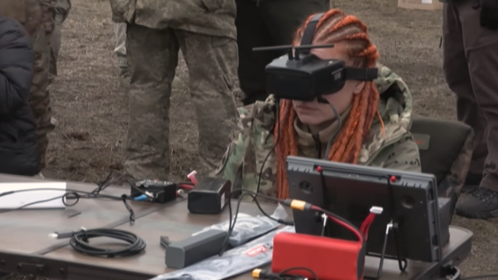 Person in military camouflage using a virtual reality headset at an outdoor defense technology setup with monitoring equipment.