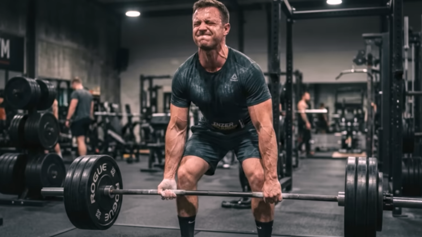 Muscular man performing a heavy deadlift in a gym while straining to lift a loaded barbell.