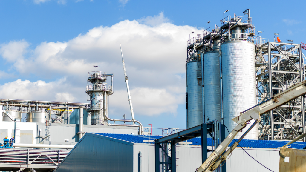 Large silver industrial storage silos and distillation towers at a modern processing plant utilizing AI in Oil Production technology under a blue sky.