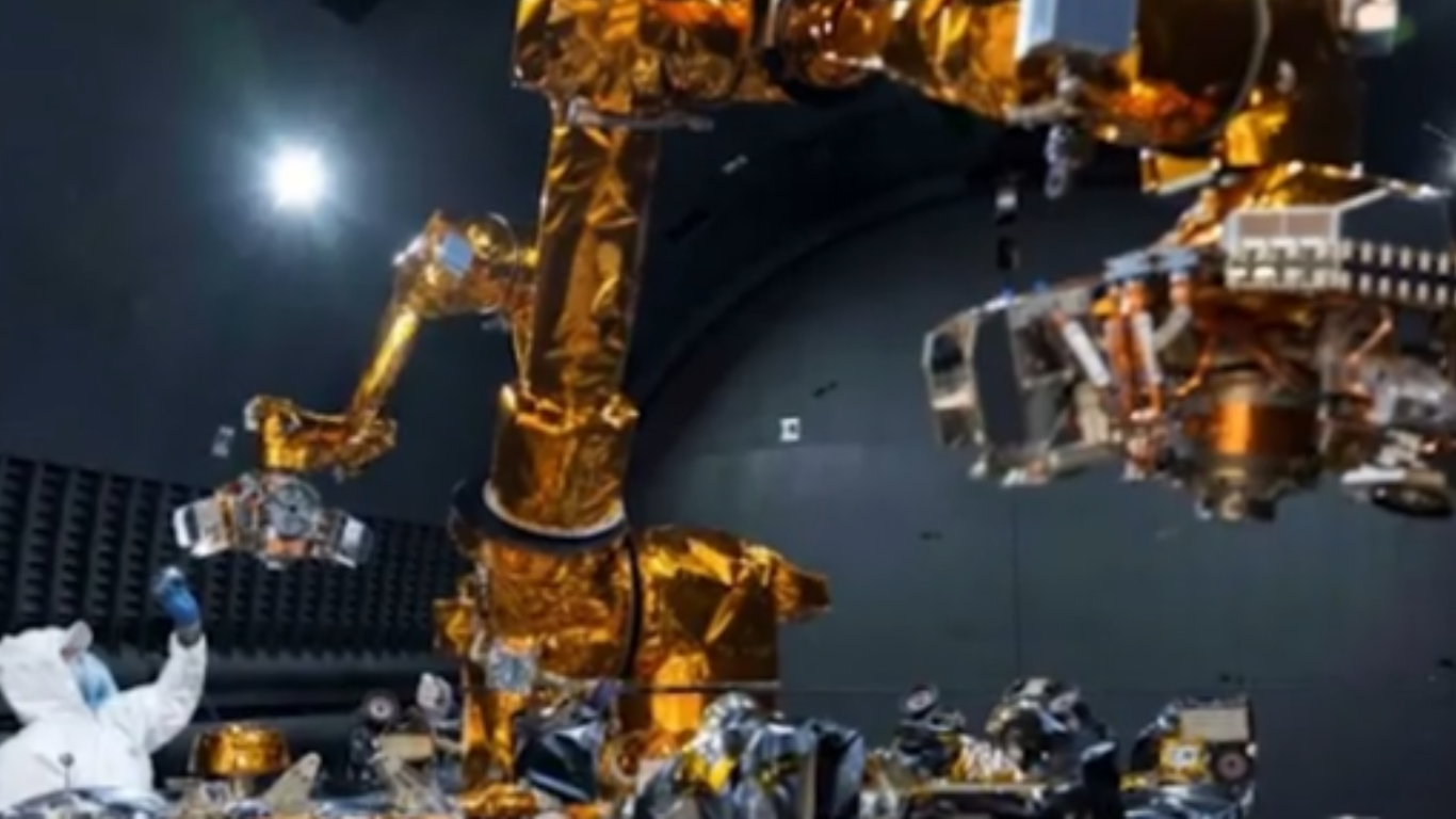 Close-up view of the golden-wrapped dual robotic arms from the Autonomous Satellite Guarding Systems project during testing inside a dark, specialized lab environment with a technician visible on the lower left. U.S. Naval Research Laboratory.