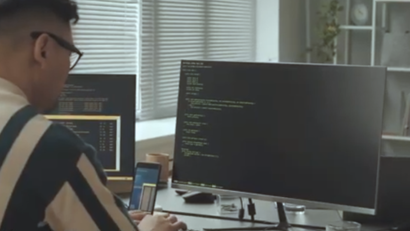 Rear view of a developer with glasses working at a desk, looking at large monitors filled with code blocks, demonstrating the development phase of Edge AI Optimization.