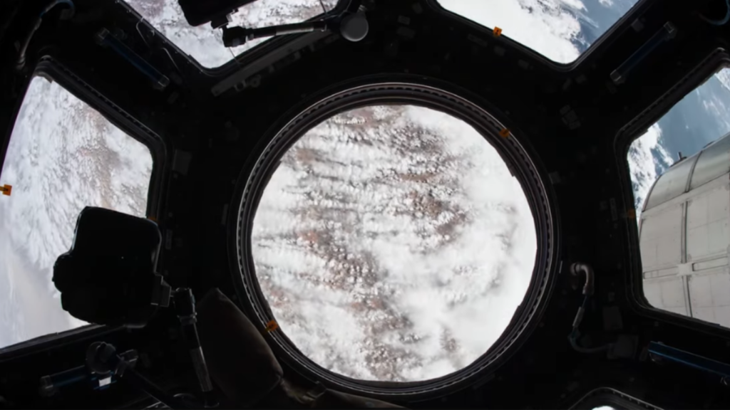 View of Earth's cloud-covered surface looking directly through a large, circular observation window inside a spacecraft, contextually framed around the concept of Space Debris as a Defensive Barrier.