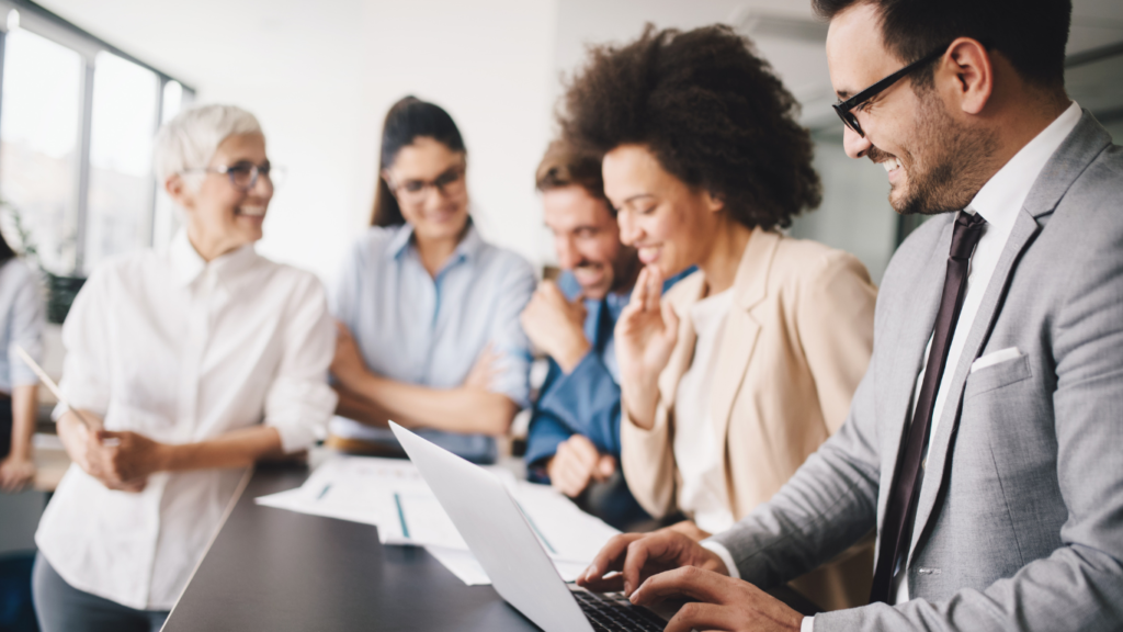 A diverse corporate team smiling and collaborating around a modern office table, featuring a businessman in a grey suit and glasses typing on a laptop in the foreground while reviewing predictive AI analytics reports with four colleagues looking on enthusiastically.
