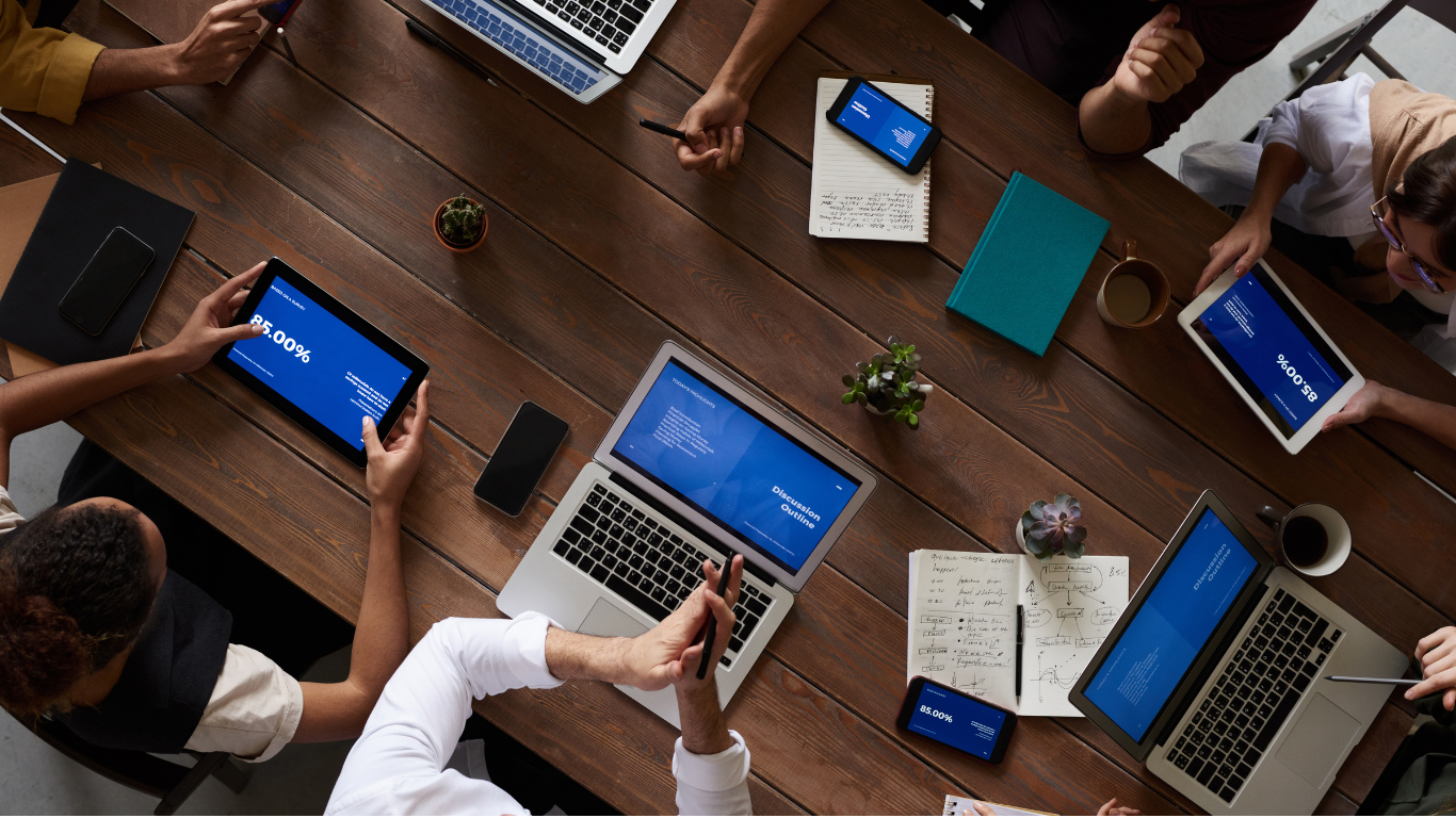 An overhead view of a diverse business team seated around a wooden conference table, collaborating with laptops, tablets, and smartphones to evaluate what key factors to consider before investing in Edge AI Hardware for Enterprise Applications.